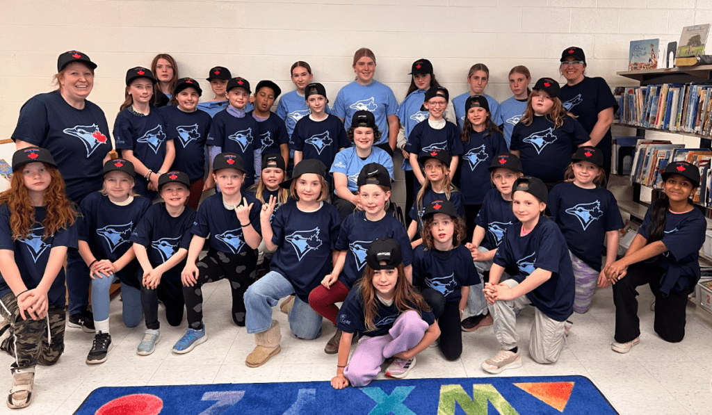 A large group photo of about two dozen children and two adult leaders posing together indoors. Everyone is wearing matching dark blue or light blue Toronto Blue Jays t-shirts and black baseball caps featuring a red maple leaf. They are smiling and posing in multiple rows, with some kneeling in the front and others standing in the back. This is in celebration of Girls at Bat.
