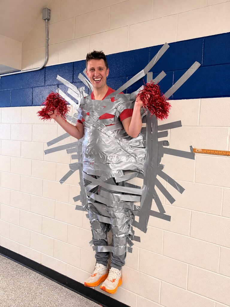 A smiling school principal, Mr. Cadeau-Mark, taped to a hallway wall with silver duct tape while holding red pom-poms as part of a Terry Fox fundraiser celebration.